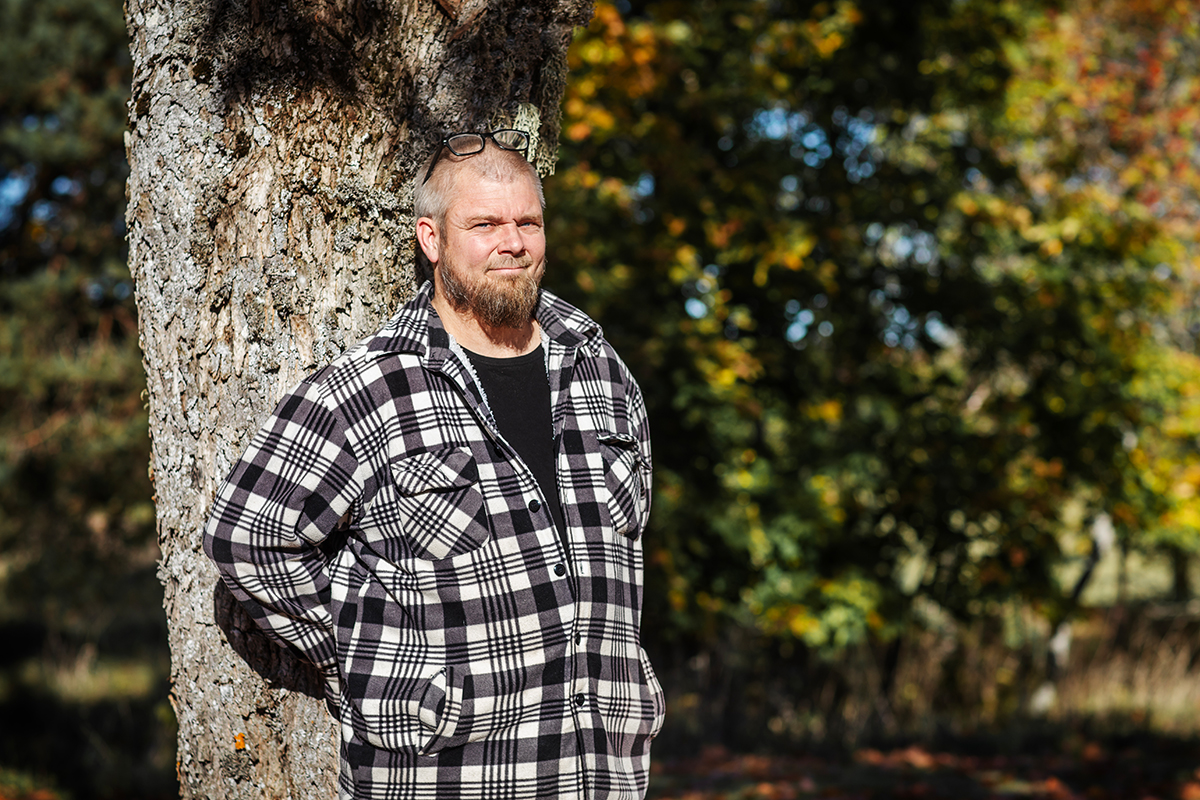 Gråhårig man med solglasögon uppskjutna i panna står lutad mot trädstam i parkmiljö.